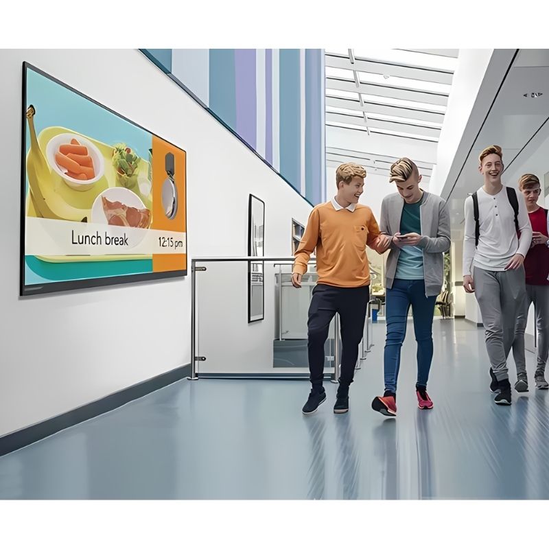 Four students walking past a Boxlight Clevertouch Max 2 interactive display mounted in a school hallway, demonstrating the CleverLive digital signage capabilities for campus communication