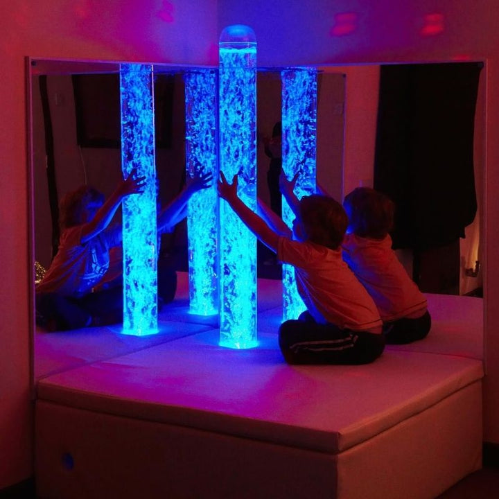 Child actively engaged with the Experia Bubble Tube Mirrors corner installation in blue light, showing the quadrupled visual experience of the shatter-resistant acrylic mirror pair