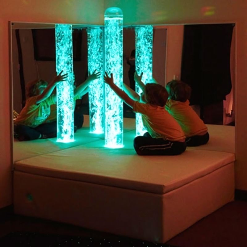 Child actively playing in front of the Experia Bubble Tube Mirrors in cyan light, showing the corner-mounted acrylic pair multiplying the bubble tube visual experience