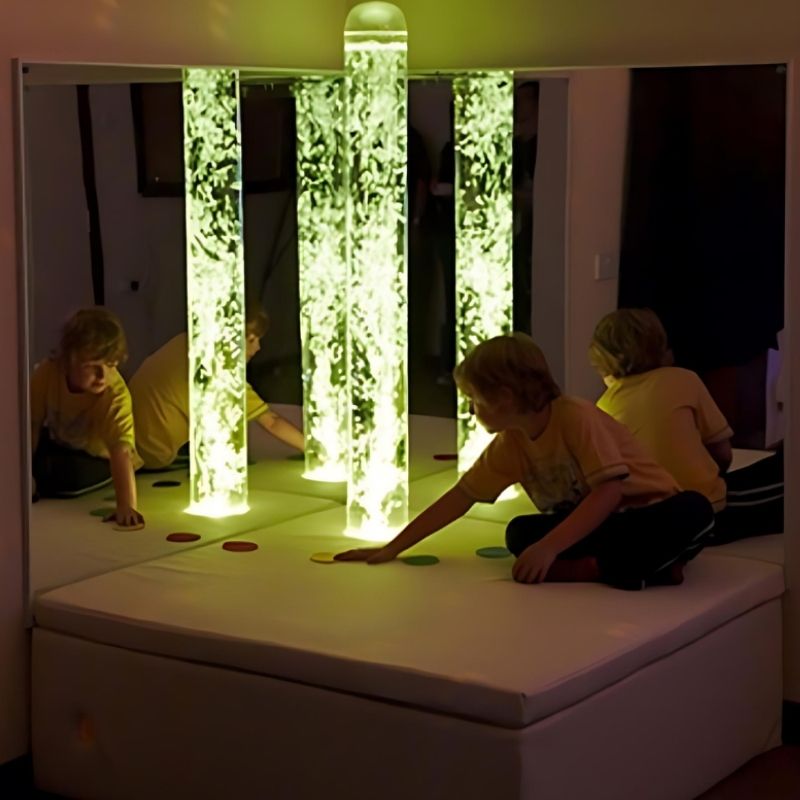 Child pressing the platform colour-switch spots on the Experia Interactive Bubble Tube Corner, showing touch-activated yellow colour control of the LED bubble tube sensory corner