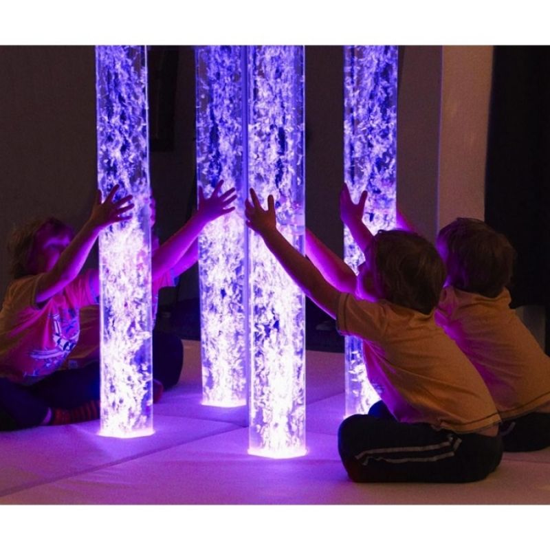 Child seated next to the Experia Touch Sensitive LED Sensory Bubble Tube engaging with the base buttons, showing the accessible touch-sensitive colour control for users with limited motor skills