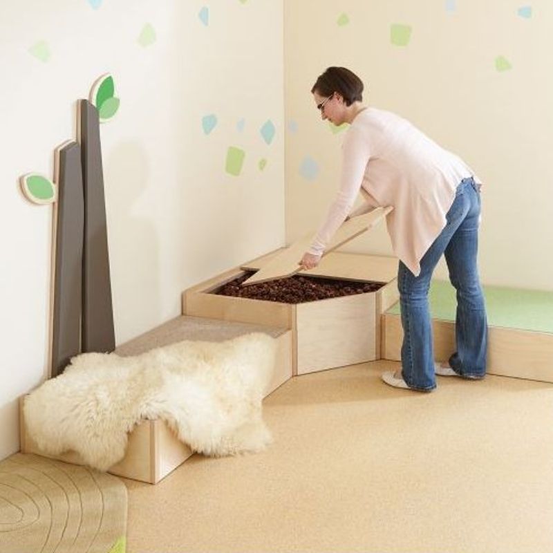 Adult opening the two-piece birch wood lid of the HABA Pro Grow.upp sensory bath platform to reveal chestnut fill, shown in a calm corner classroom setting