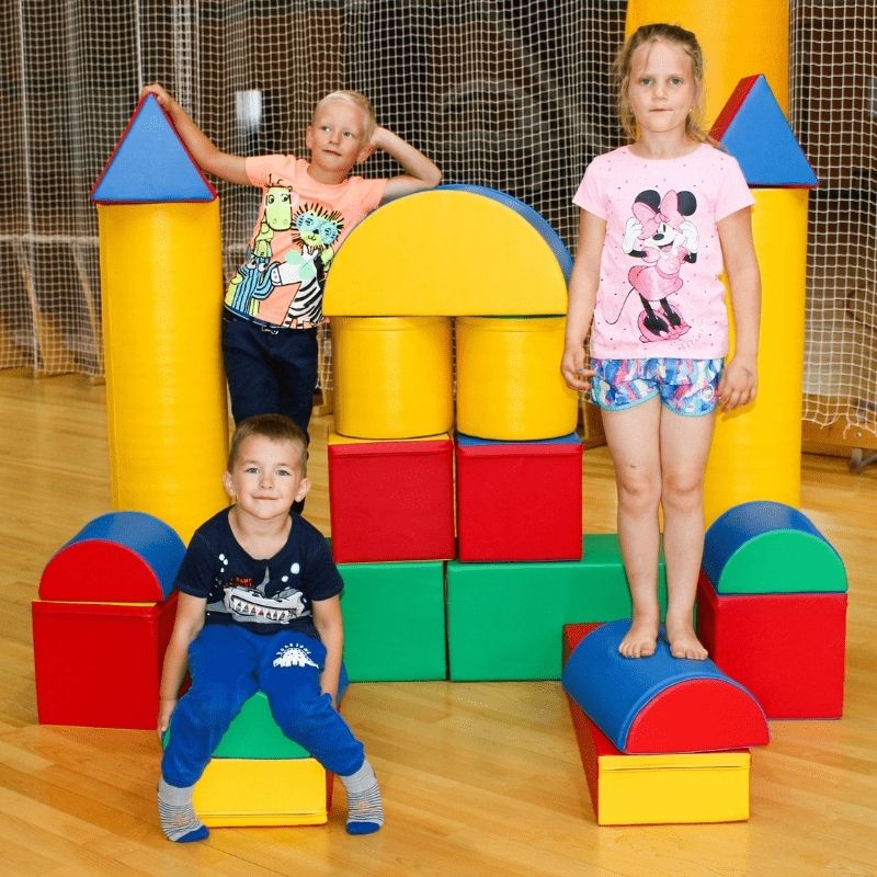 Three children building and climbing the IGLU Soft Play Fortress Set in multi-colour configuration, demonstrating imaginative play and gross motor development