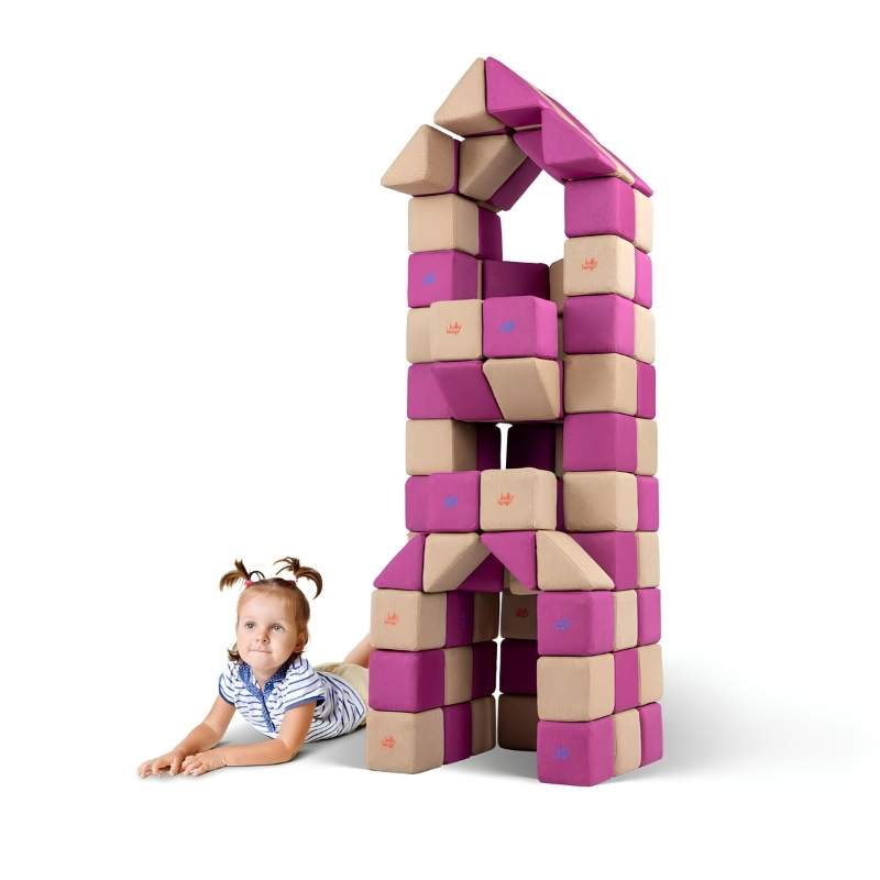A girl lies beside a tall tower structure built from JollyHeap Basic Set magnetic foam blocks, showing the vertical building possibilities of this soft play set.
