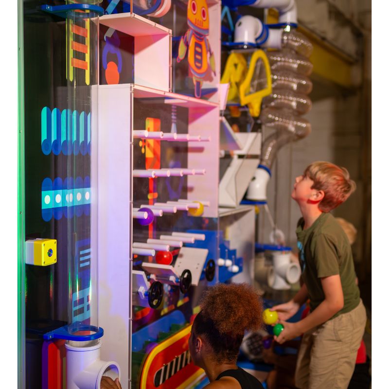 Two children of different ages exploring the tall panel section of the LiteZilla Ultimate Ball Launcher, with one child holding a ball beside the clear lit tube and plinko elements against a custom robot-themed mural backdrop