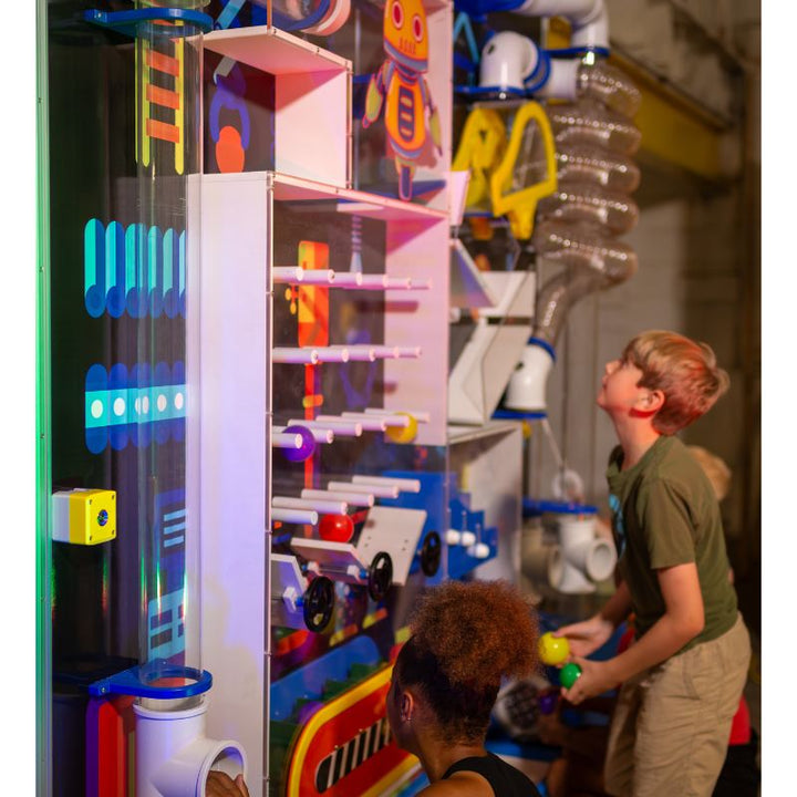 Two children of different ages exploring the tall panel section of the LiteZilla Ultimate Ball Launcher, with one child holding a ball beside the clear lit tube and plinko elements against a custom robot-themed mural backdrop