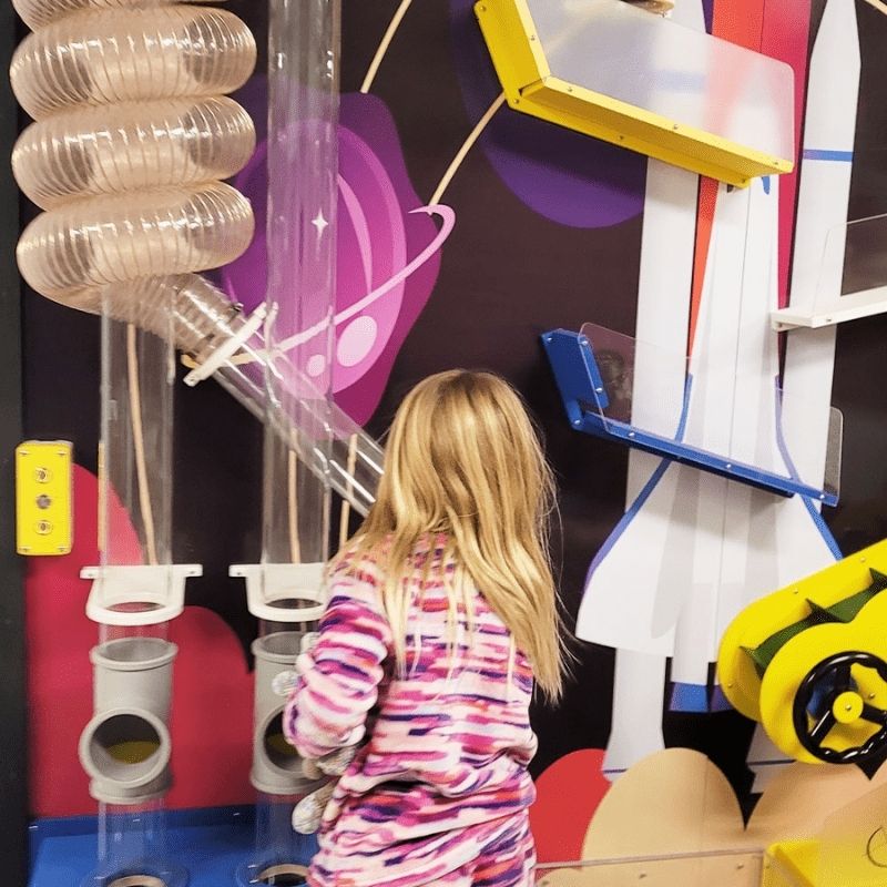 A child exploring the clear spiral tubing and pipe junction components of the LiteZilla Ultimate Ball Launcher, shown with a custom space-themed mural backdrop as an example of the exhibit's fully customizable design