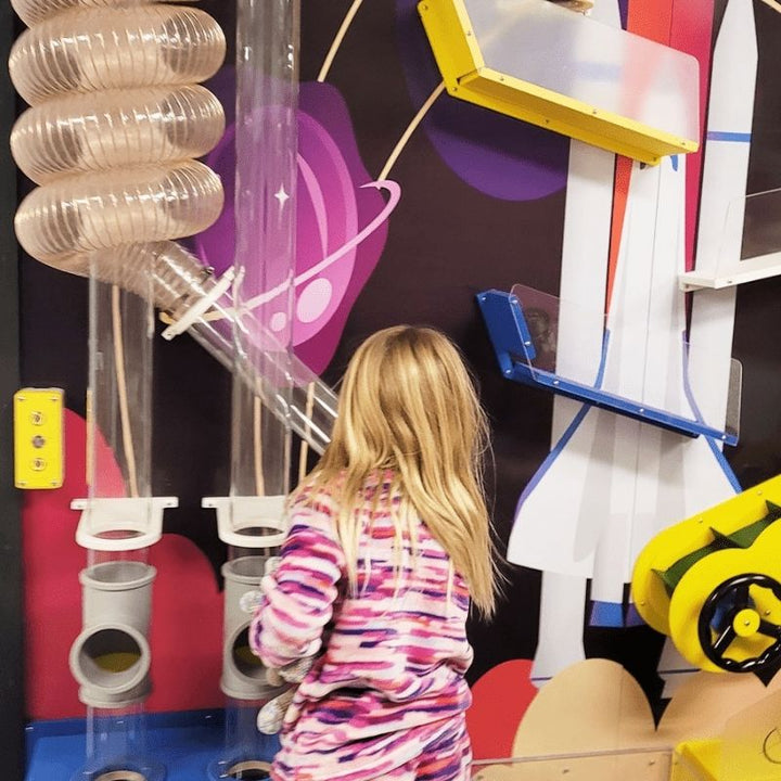 A child exploring the clear spiral tubing and pipe junction components of the LiteZilla Ultimate Ball Launcher, shown with a custom space-themed mural backdrop as an example of the exhibit's fully customizable design