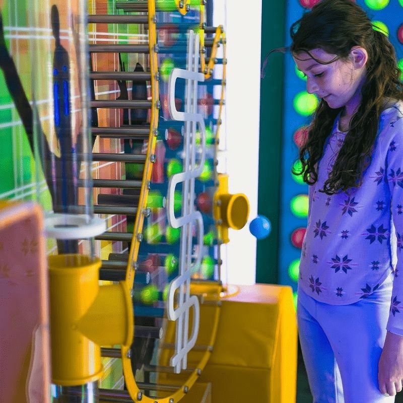 A child watching balls travel through the chain conveyor pathway of the LiteZilla Ultimate Ball Launcher, capturing the focused cause-and-effect engagement the STEM sensory exhibit delivers