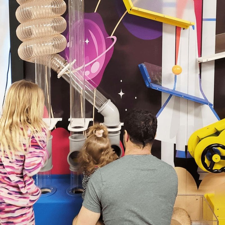 A child and adult exploring the LiteZilla Ultimate Ball Launcher together, examining clear tube junctions and air-powered pathways against a custom space-themed mural backdrop