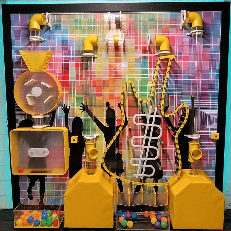 Full front view of the LiteZilla Ultimate Ball Launcher exhibit showing the complete air-powered tube network, spinning disc, chain conveyor, and plinko pathway mounted against a customizable colourful mural backdrop