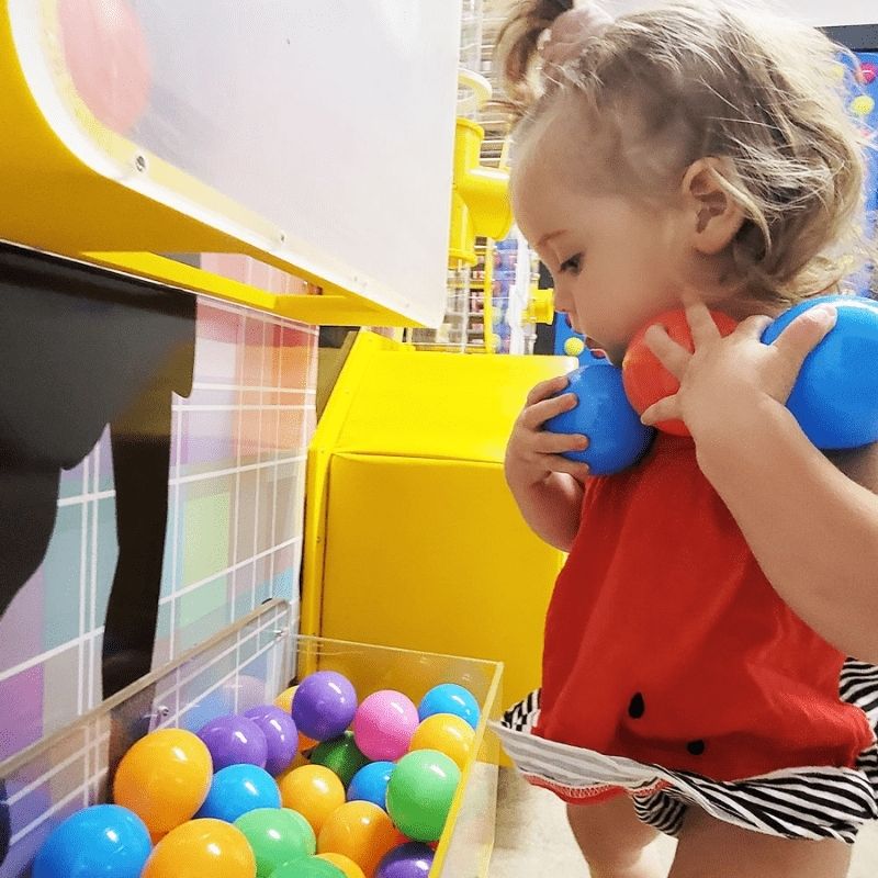 A toddler holding a ball and exploring the launch unit of the LiteZilla Ultimate Ball Launcher air-powered sensory exhibit, with colourful balls visible in the collection tray