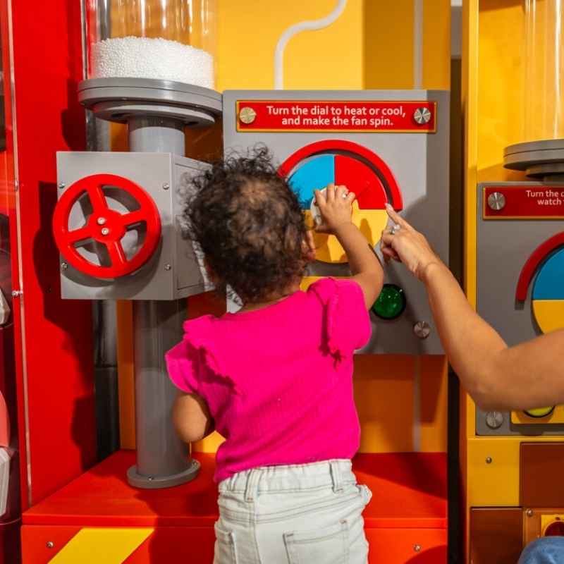 A young child turning the control wheel on the LiteZilla Hurricane Tube cause and effect wall exhibit, with an adult guiding the interaction