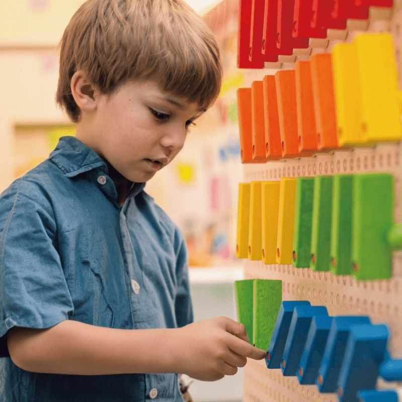 Child arranging rainbow-coloured domino tiles on the LiteZilla KidWorx wall-mounted STEM panel, developing sequencing and fine motor skills through open-ended play