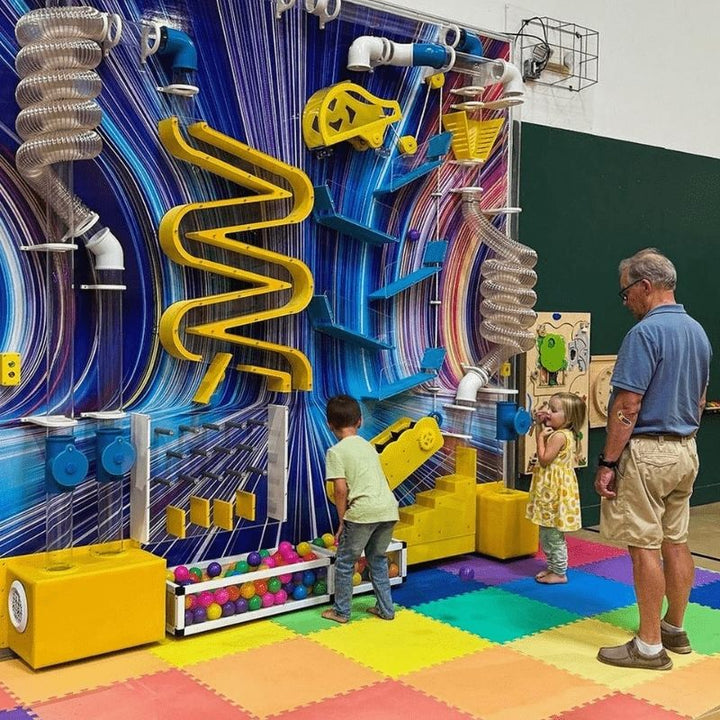 LiteZilla Ultimate Ball Launcher installed in a children's play space, showing two young children and an adult watching balls travel through clear tubing, yellow gear tracks, and plinko pathways of this air-powered STEM sensory exhibit