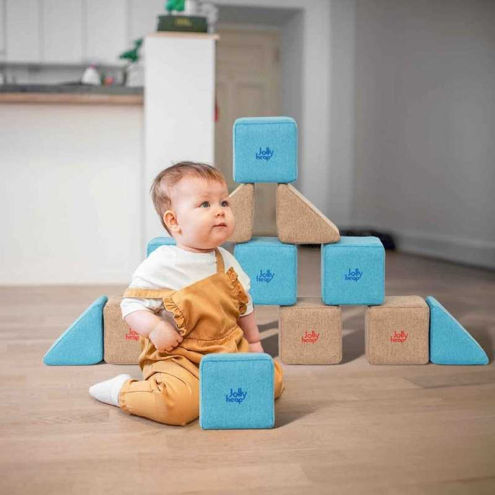 Baby playing beside a pyramid shaped structure built from JollyHeap Pixi soft magnetic building blocks in teal and tan