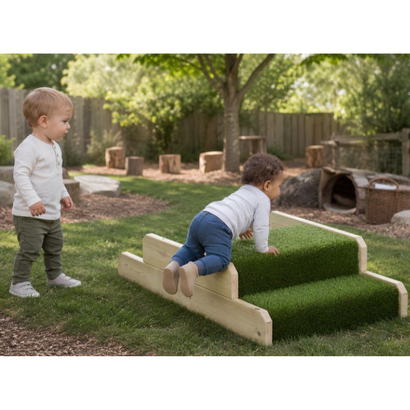 A toddler climbing the turf-covered steps of the ONP Infant Step and Ramp outdoors, while another child watches nearby