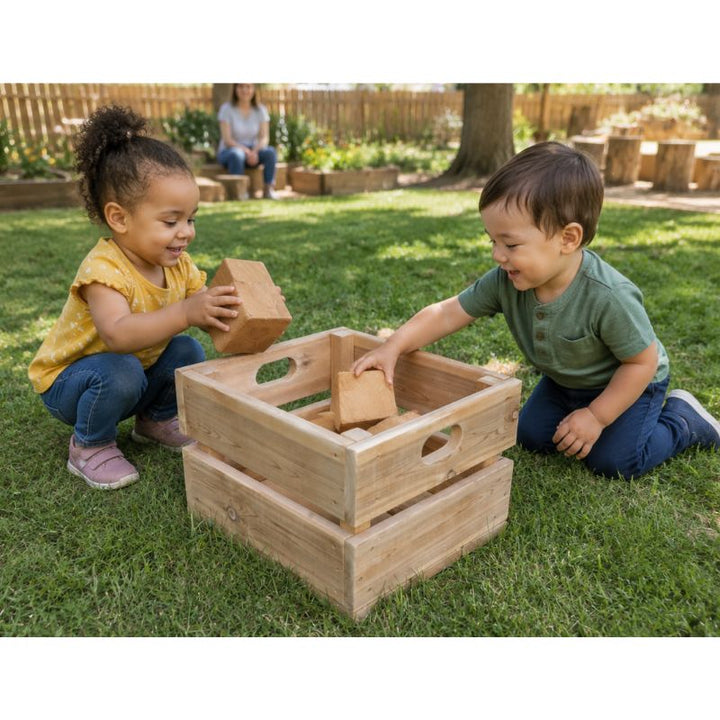 Two children placing wooden blocks into the ONP Large Outdoor Box outdoors, a storage crate with cut-out handles for easy carrying