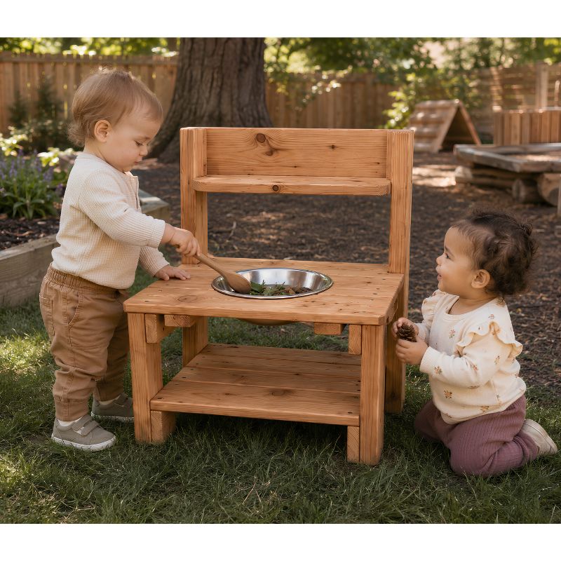 Two toddlers playing at the ONP Mini Mud Kitchen outdoors, stirring leaves in the stainless steel bowl with a wooden spoon