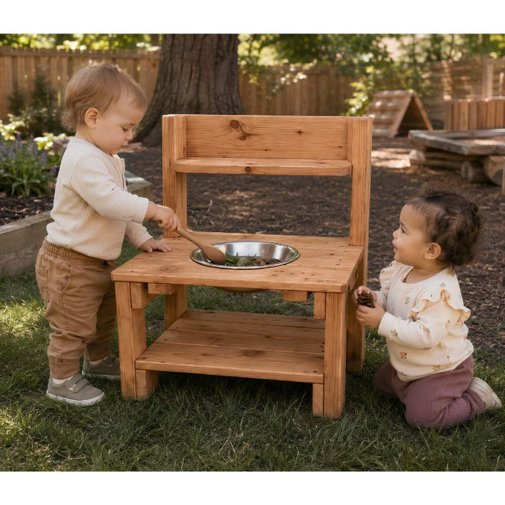 Two toddlers playing at the ONP Mini Mud Kitchen outdoors, stirring leaves in the stainless steel bowl with a wooden spoon