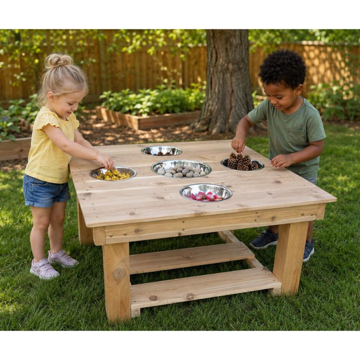 Two children playing at the ONP Outdoor Messy Activity Table on grass, sorting flower petals, stones and pinecones into the five built-in bowls