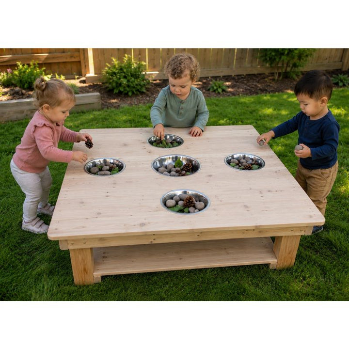 Three children exploring the ONP Outdoor Messy Activity Table outdoors, dropping stones and pinecones into the five stainless steel bowls