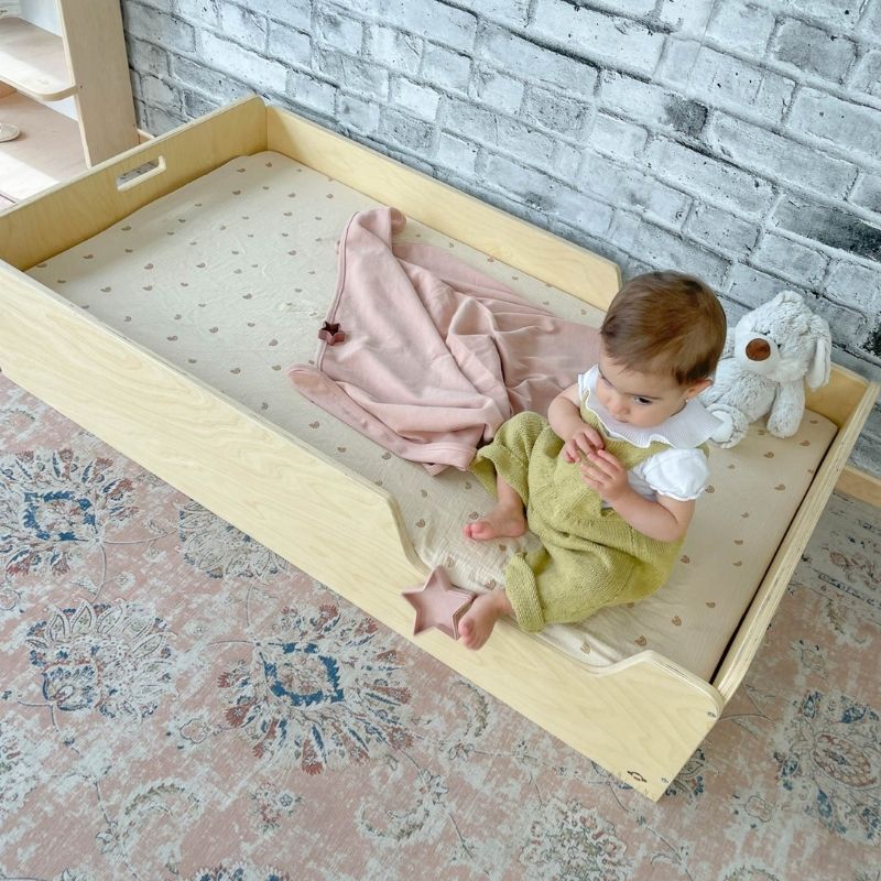Top-down view of a child in the Sapiens Child Apple Montessori floor bed with rails, showing the full sleeping surface, protective borders, and adjustable side shelf