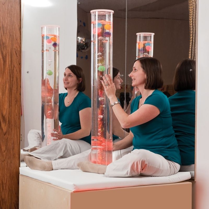 Woman seated on the padded platform of a Southpaw sensory bubble tube with ping pong insert, touching the tube in an indoor therapy room setting