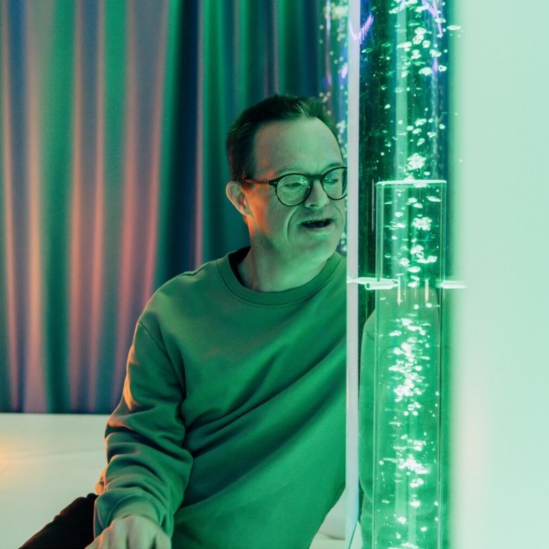 Adult alongside TFH Aquarium Bubble Tube 6" in green LED mode demonstrating the calming bubble column for adult sensory use in care homes and OT clinics