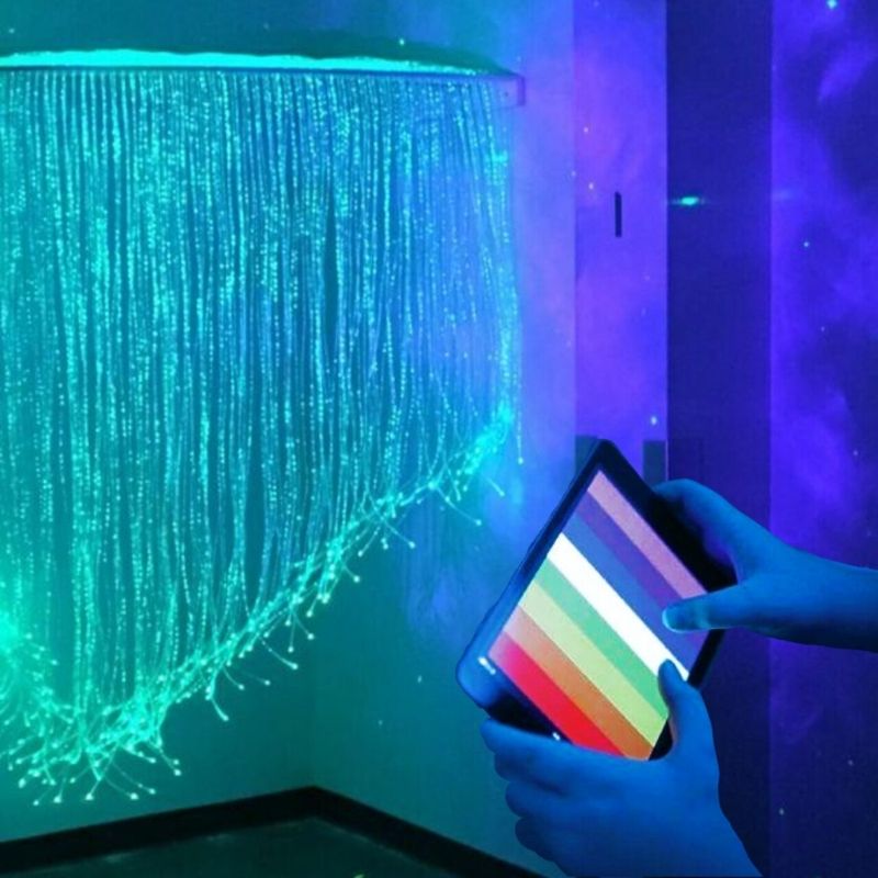 TFH Genie Quarter Circle Fibre Optic Curtain installed in a sensory room corner with a user holding the colour controller, showing the cascading fibre optic tails and MSE Genie colour control in use