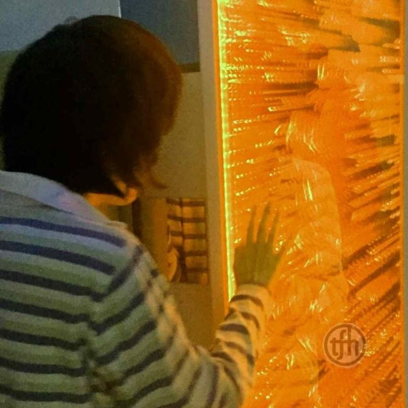 Back view of a child touching the TFH Infinity Bubble Wall lit in orange, showing tactile and visual sensory stimulation through direct wall contact