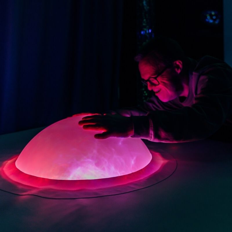 Adult interacting with the TFH Interactive Dome Swirl Podium in pink light, showing the sound-activated colour response of the sensory light dome in a therapy room setting
