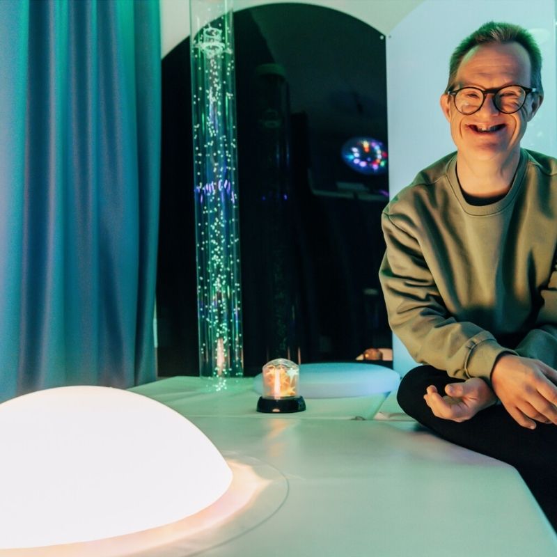 Happy adult beside the TFH Interactive Dome Swirl Podium in a sensory room, demonstrating the eye-level podium mount for up-close personal sound-activated light interaction