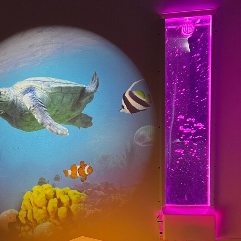 TFH Lights & Bubbles Water Panel glowing in a darkened sensory room alongside a projector, showing the wall-mounted bubble wall with colour-cycling LED display