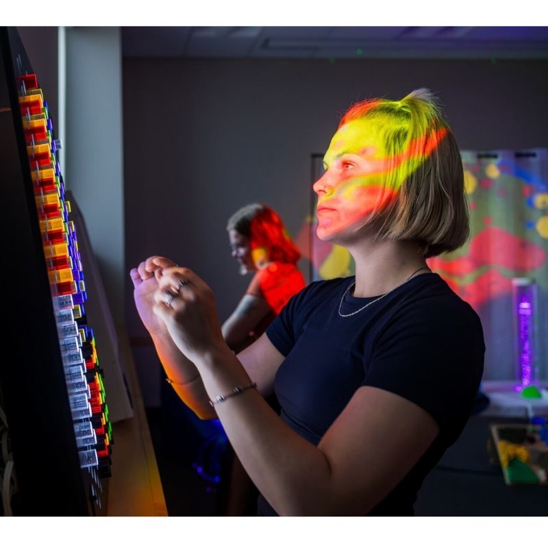 TFH Rod Wall Activity Panel with an adult woman engaging from the side, demonstrating the wall-mounted illuminated peg panel for occupational therapy and sensory room use