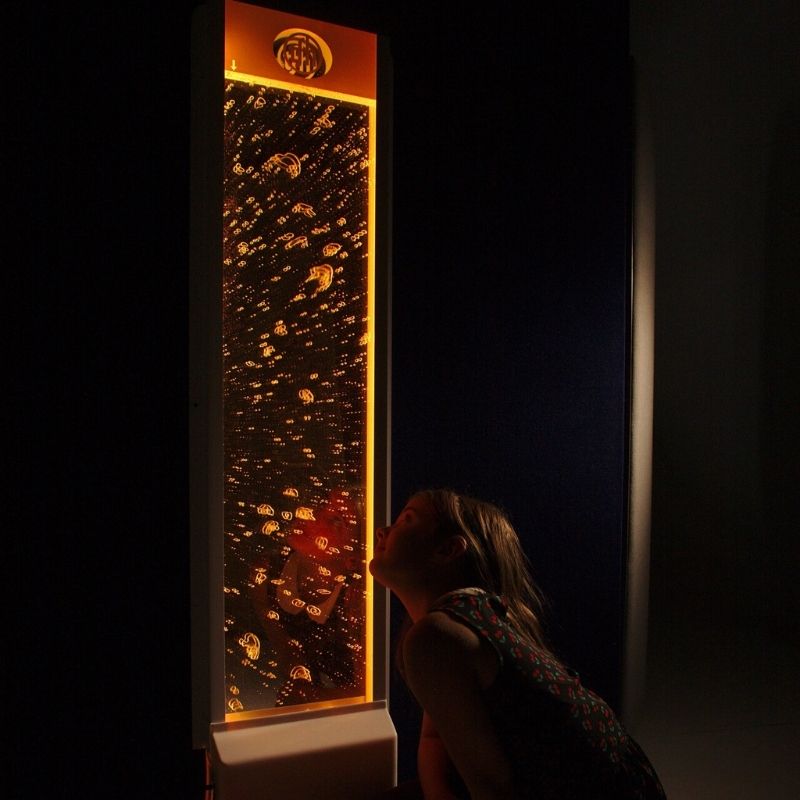 Child engaging with the TFH Lights & Bubbles Water Panel in a dark room setting with orange LED light, showing the calming bubble wall effect