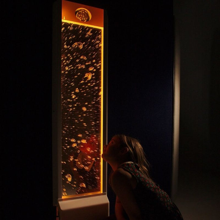 Child engaging with the TFH Lights & Bubbles Water Panel in a dark room setting with orange LED light, showing the calming bubble wall effect