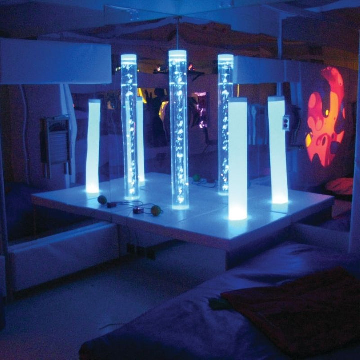 TFH Square Podium Base in a darkened sensory room with bubble tube glowing in blue light, showing the mirrored acrylic sides amplifying the colour-changing sensory environment