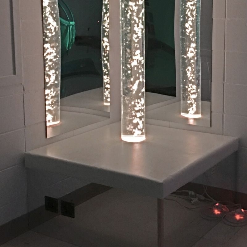 TFH Square Podium Base in a sensory room landscape view with the bubble tube glowing in warm light, showing the mirrored sides reflecting colour across the sensory room floor