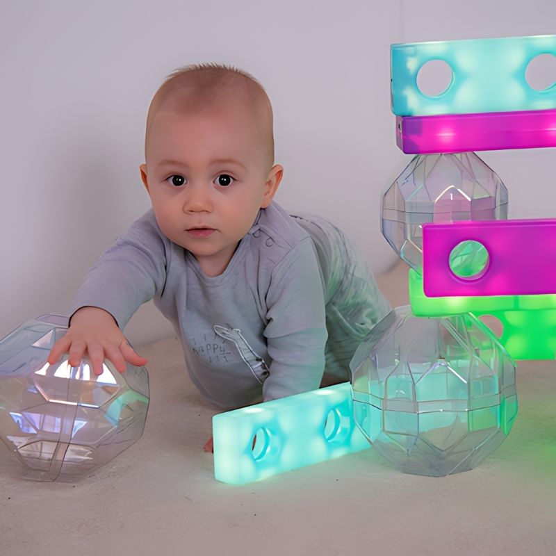 Baby reaching toward TTS Stack and Build Glow Blocks stacked in pink, green and teal alongside a clear sensory sphere, showing early sensory exploration and visual stimulation for infants