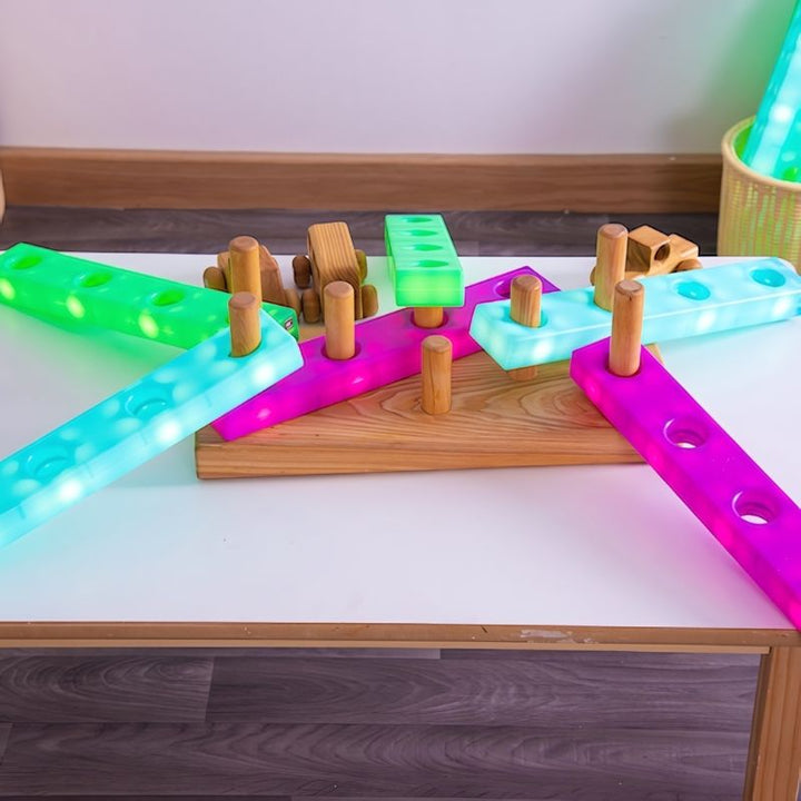 TTS Stack and Build Glow Blocks in blue, green and pink arranged on a light table with wooden figures, demonstrating open-ended construction and sensory light play