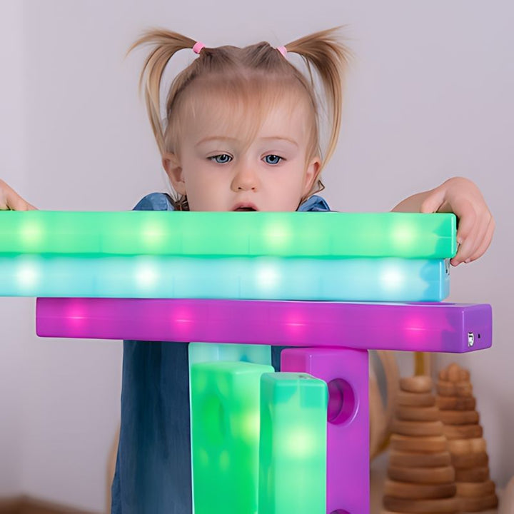 Toddler examining a stacked tower of TTS Stack and Build Glow Blocks in green, pink and teal, exploring the LED glow up close and developing visual attention and fine motor stacking skills