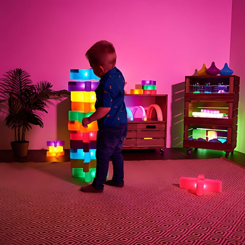 Young child stacking a tall illuminated tower of TTS Glow Crosses and light-up bricks in a sensory play room, demonstrating vertical construction play and balance skills