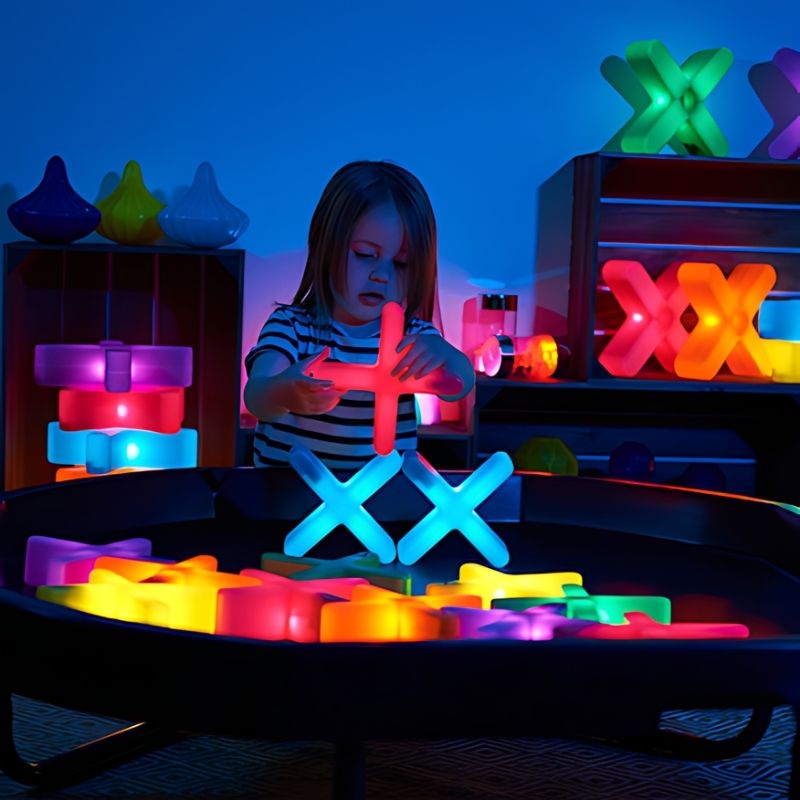 Girl building with TTS Glow Crosses on a dark activity tray surrounded by a full range of illuminated sensory construction resources, supporting loose parts play and visual sensory engagement