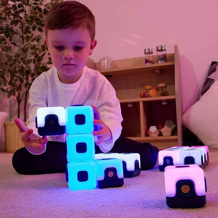 Young boy building with TTS Glow Sequencing Cubes in cyan and white on a carpeted floor at home, with a connected magnetic row of purple cubes alongside, showing independent sensory construction play