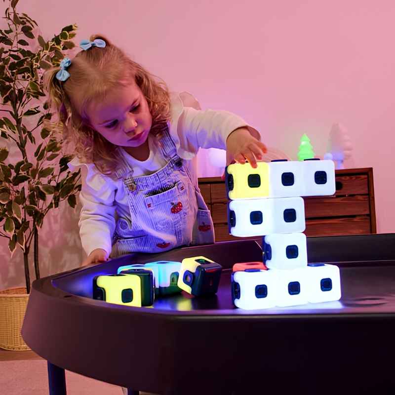 Young child building a tall stack of TTS Glow Sequencing Cubes on a tray table, with loose cubes glowing in multiple colours nearby, showing open-ended construction play