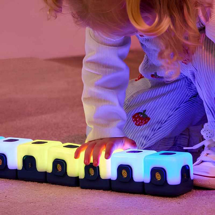 Close-up of a child's hand pressing TTS Glow Sequencing Cubes linked in a horizontal chain, showing the tap-to-change colour mechanic and magnetic connection