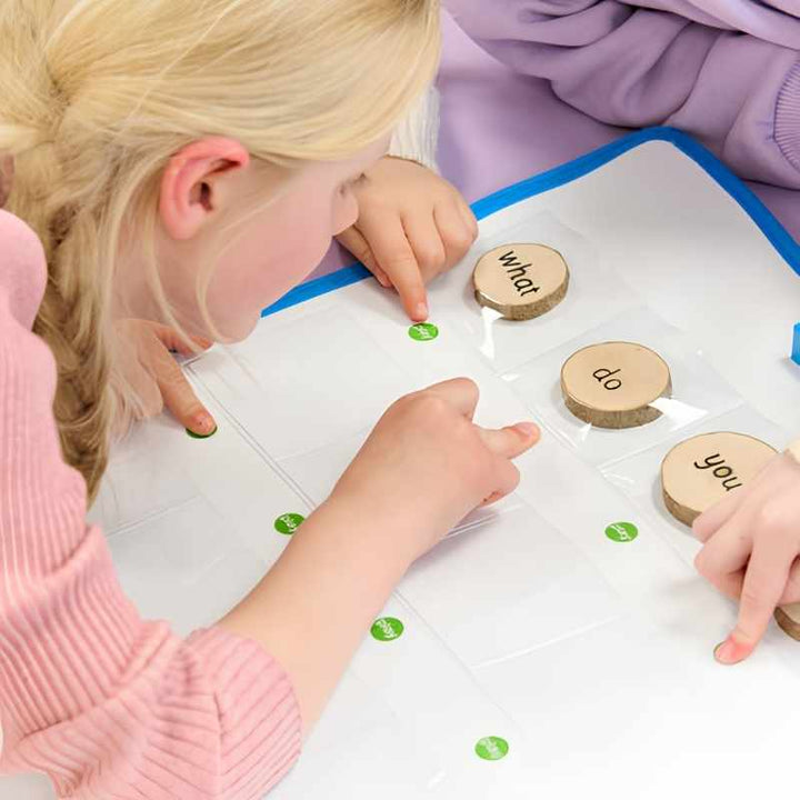 A young child pressing a green play button on the TTS Interactive Talking Mat on a table with wooden word tokens in the pockets, demonstrating independent use for oral language development