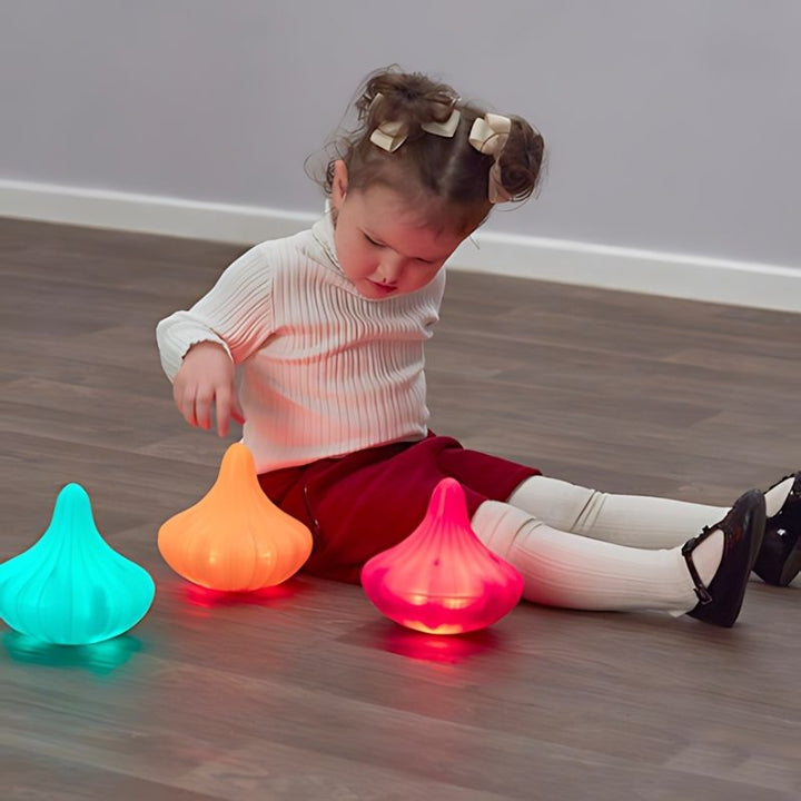 Young child reaching toward three TTS Light Up Twist and Turn Spinning Tops glowing in teal, orange, and red, illustrating open-ended sensory and investigative play for early years settings