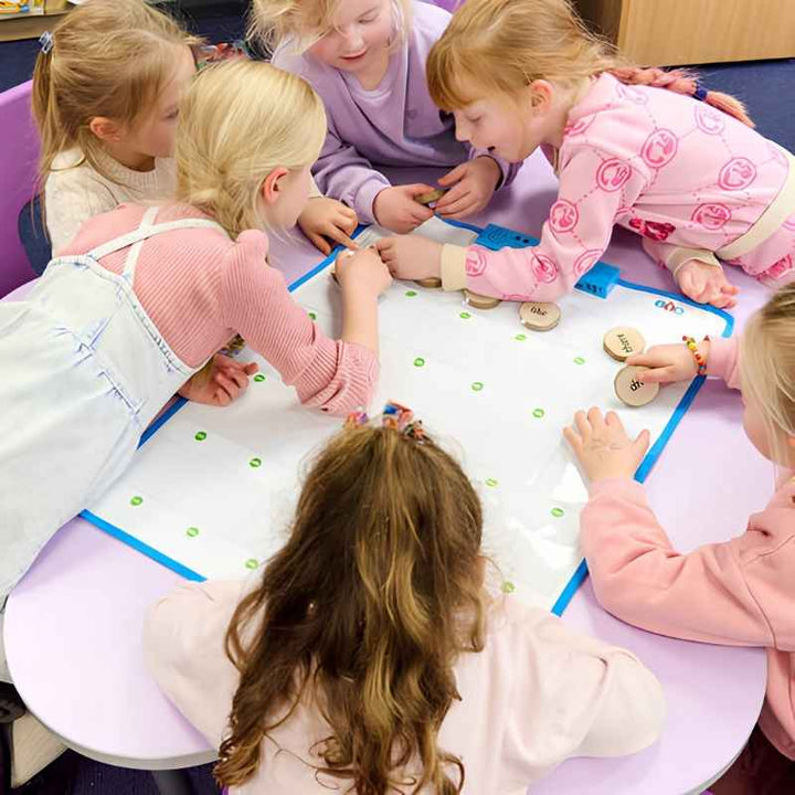 A group of six young children collaborating around the TTS Interactive Talking Mat during a classroom speaking and listening activity using wooden word tokens